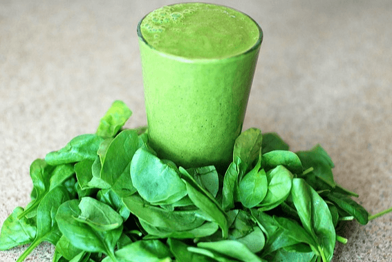 시금치를 간 녹색의 시금치 주스가 유리컵 가득 담겨있고, 컵 앞에는 시금치 잎들이 가득하다.