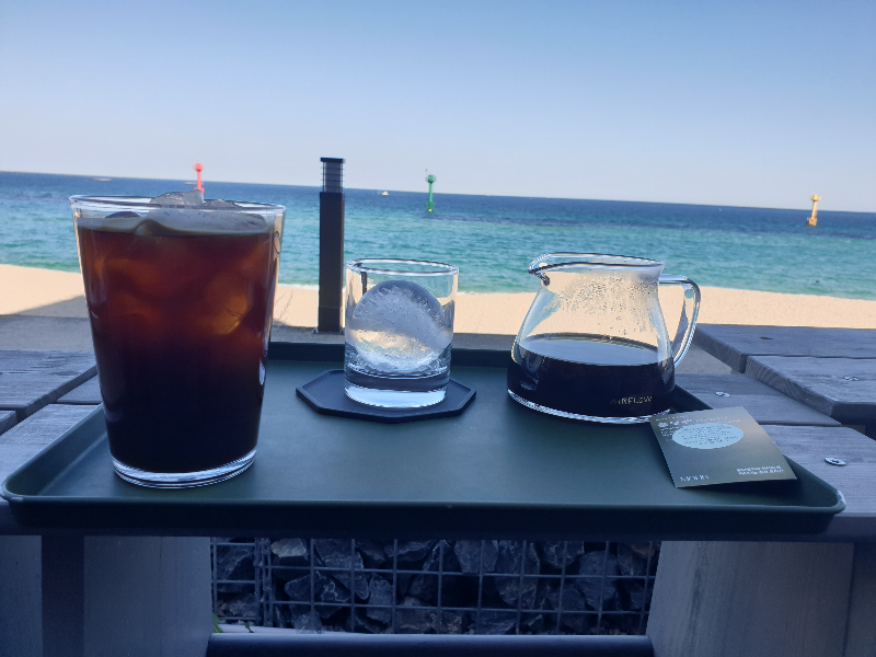바다를 배경으로 아이스 아메리카노와 브루잉 그리고 얼음이 담킨 컵이 놓여 있다.