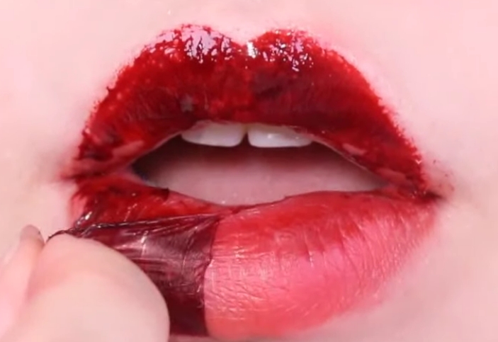 입술에 빨간 타투 립스틱을 바르고 겉의 마른 스티커 부분을 손가락으로 벗겨내는 모습입니다.