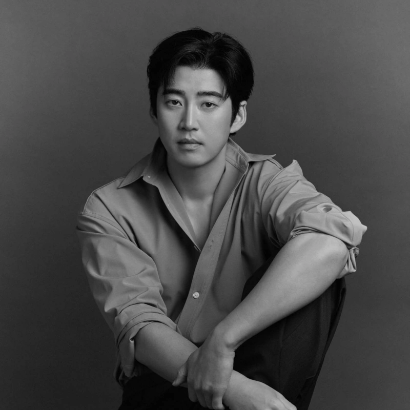 흑백의 회색 배경에 검은 머리와 와이셔츠를 입고 의자에 앉아 있는 배우 윤계상