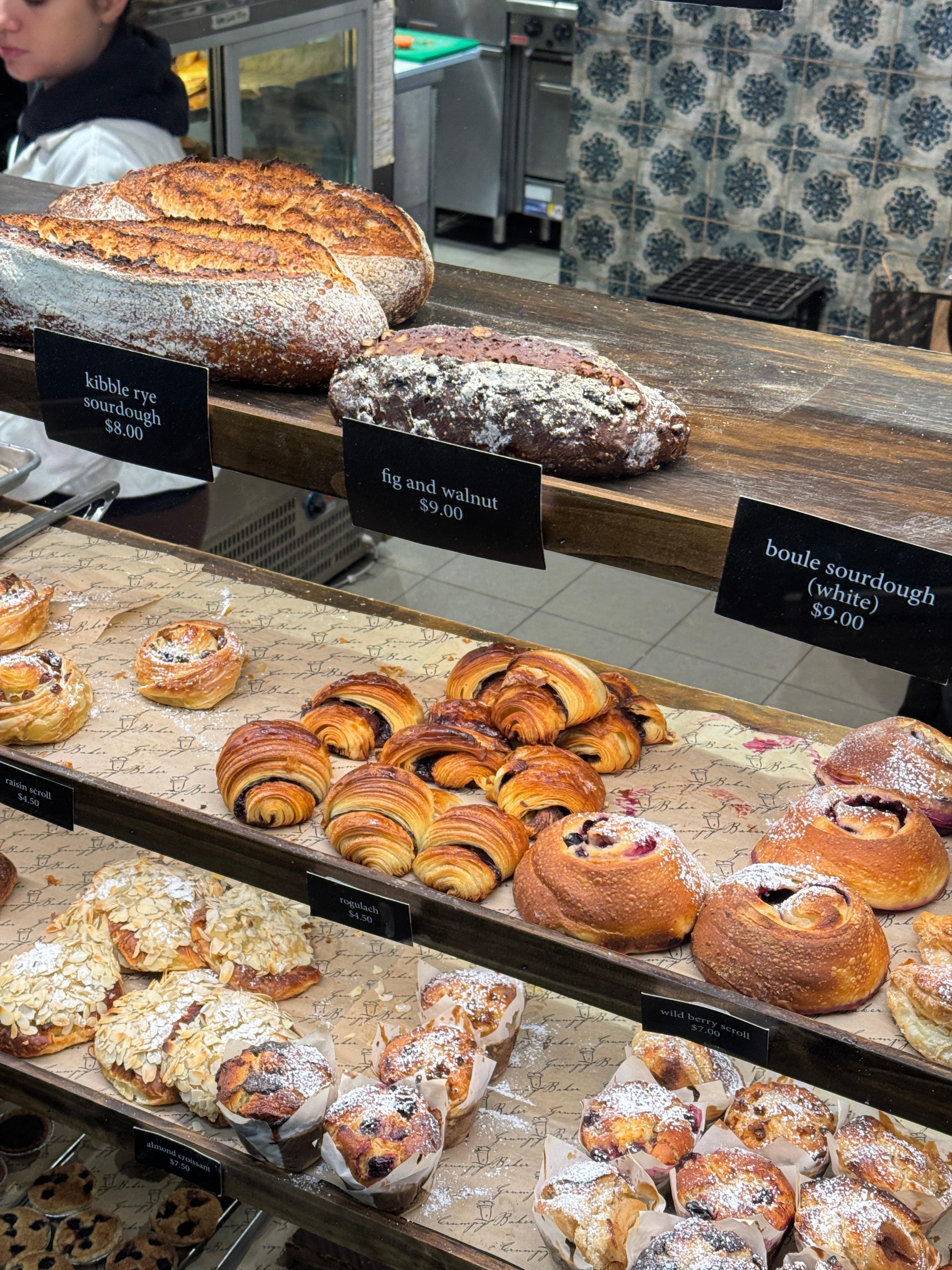 The Grumpy Baker 빵 진열대 클로즈업 – Kibble Rye 사워도우, Fig and Walnut, 초코 크루아상, Wild Berry Scroll, Almond Croissant 등 장인 빵 근접 샷