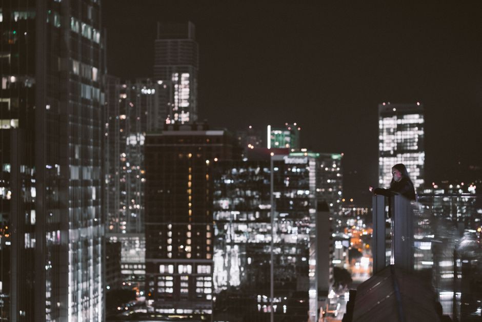 A person gazes over a bustling cityscape from a rooftop at night, surrounded by illuminated skyscrapers.