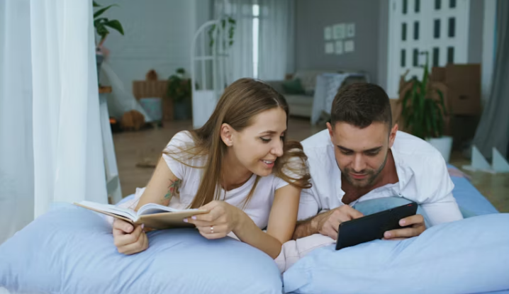 침대 위에서 함께 책과 태블릿을 보며 정보를 나누는 젊은 맞벌이 부부의 모습