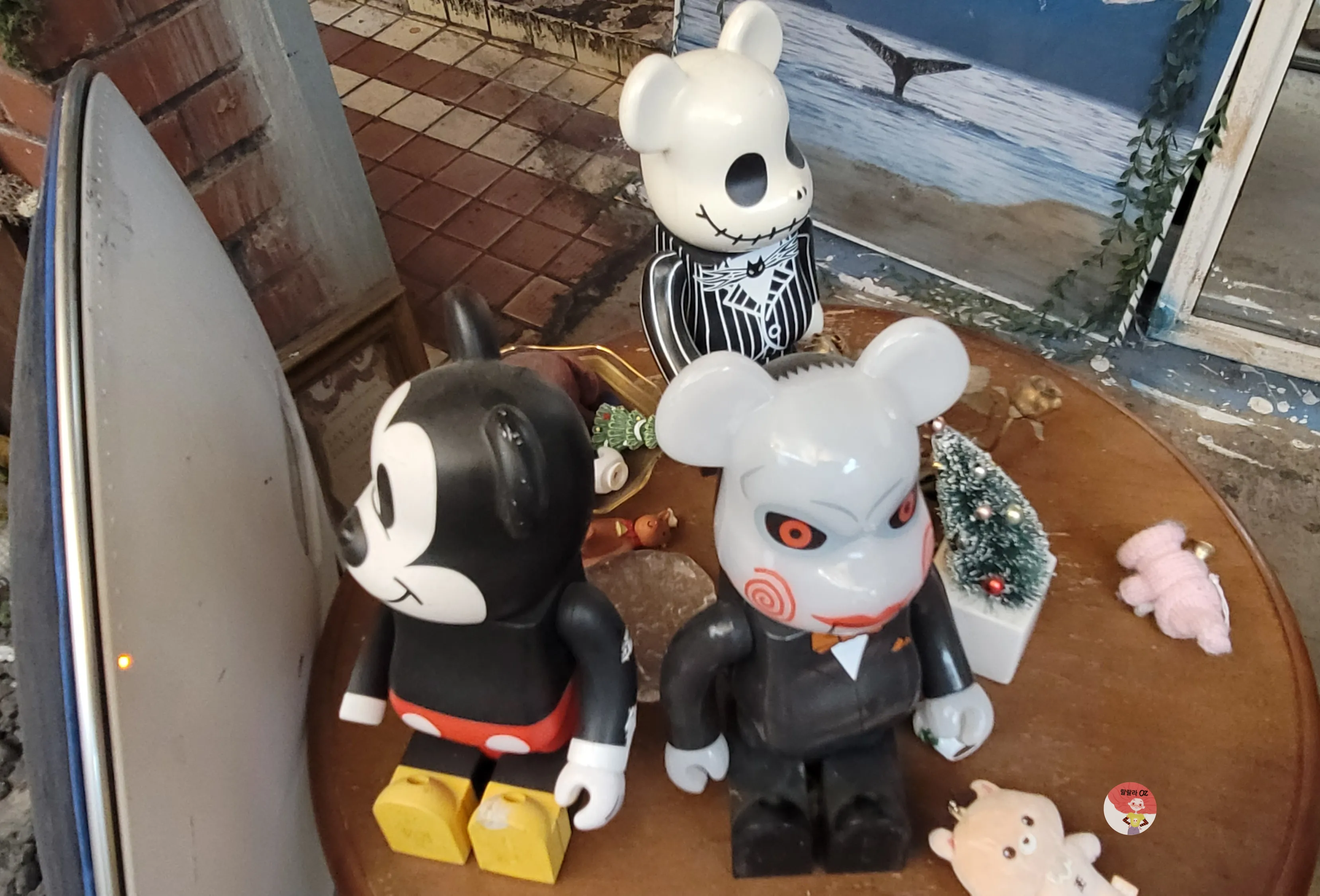 둥근 나무 테이블 위에 세 개의 곰 모양 피규어와 작은 장식품들이 놓여 있음. 검은색 캐릭터, 회색 공포 테마 피규어, 흰색 해골 무늬 피규어가 있으며 주변에는 미니 트리와 작은 인형들이 보임.
