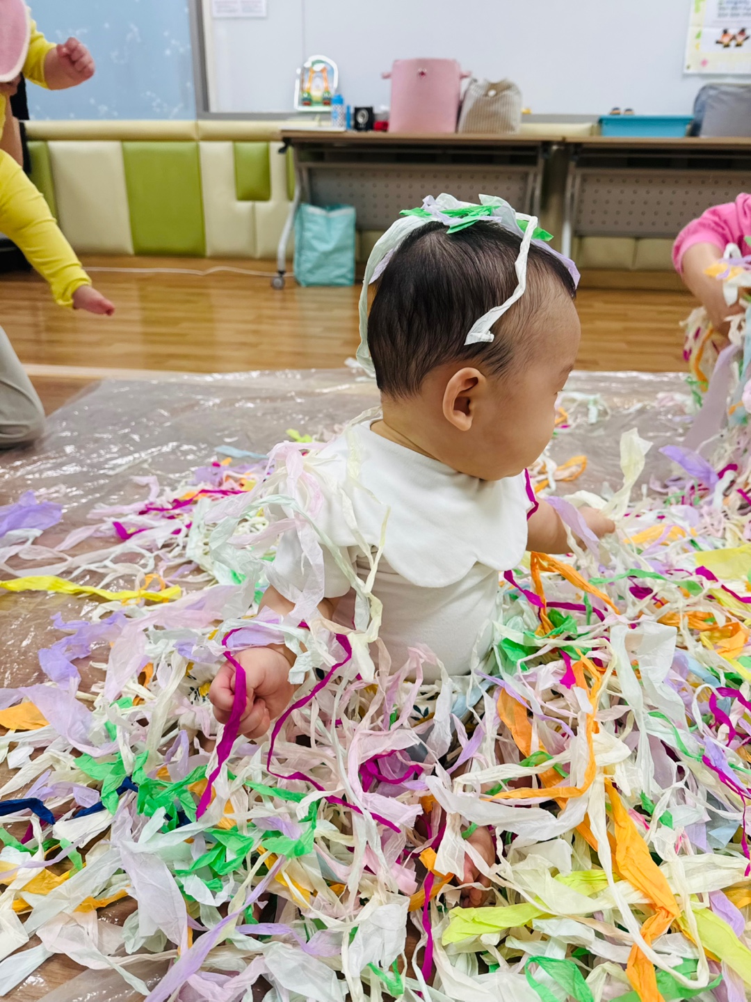 하얀 옷을 입고 색색의 습자지 위에 앉아 옆을 바라보는 9개월 아기