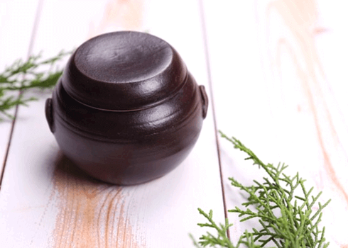 나무 바닥에 작은 된장 항아리가 있다. 항아리는 고동색의 도자기 형태이며, 주변에는 녹색의 나뭇잎이 있다.