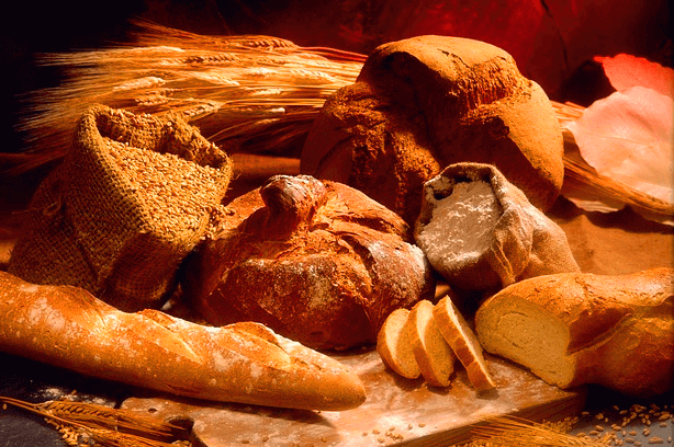 커다란 빵들이 가득 모여있다. 기다란 바게트 빵, 둥그런 통밀빵과 밀가루가 담긴 포대, 옥수수가 담긴 포대, 통밀곡식이 있다.