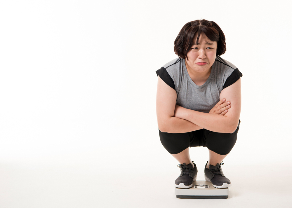 한 여성이 체중계 위에 올라가 쪼그려 앉아 있는 모습입니다. 그녀는 회색 운동복을 입고 있으며, 팔짱을 낀 채 입을 삐죽 내밀며 불만스러운 표정을 짓고 있습니다. 다이어트나 체중 증가에 대한 고민을 표현하는 장면으로 보입니다.