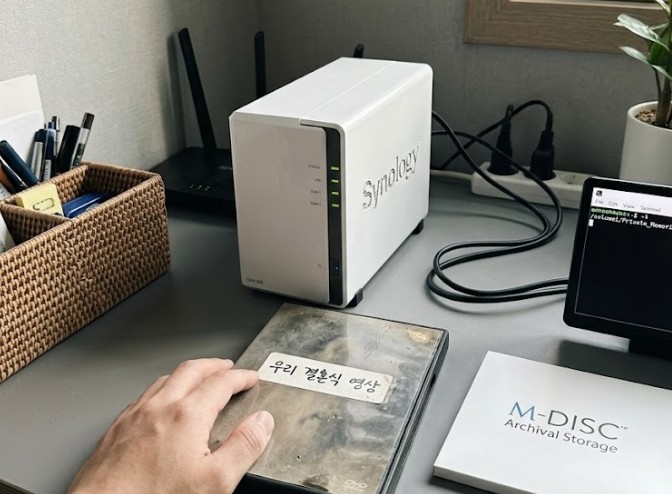 집 거실 책상 위 하얀색 Synology NAS와 그 옆에 놓인 먼지 쌓인 옛날 '우리 결혼식 영상' DVD 케이스. 부모 중 한 명의 손이 그 케이스를 만지고 있으며, 옆에는 최신 M-DISC 한 장이 놓여 있다. 포스팅에서 다룬 '가정용 NAS 수명 고민'과 '현실적 데이터 보존법'을 시각화한 현실적인 오리지널 이미지.