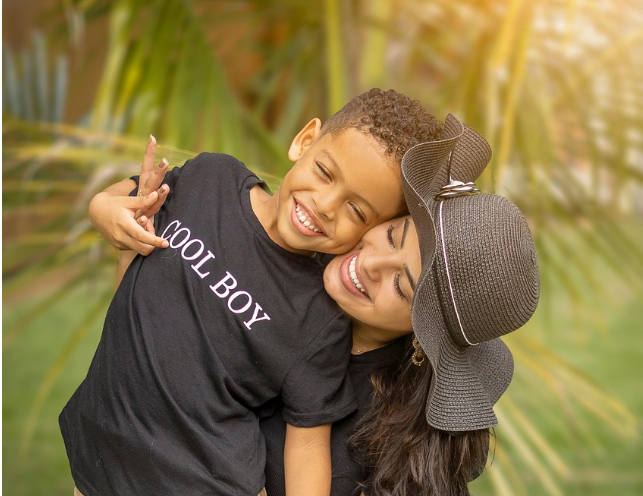 밝게 웃는 어린 아들과 미소 짓는 엄마가 함께 포옹하는 따뜻한 장면. 야외 자연 배경 속에서 정서적 안정감과 가족 간의 유대감을 표현하고 있음.