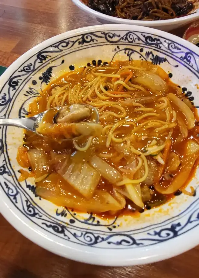 보배반점 - 나무 테이블위 흰색 바탕 파란 줄무늬 야채와 해산물이 가득 들어있는 주황색 짬뽕의 해산물이 숟가락에 담긴 이미지