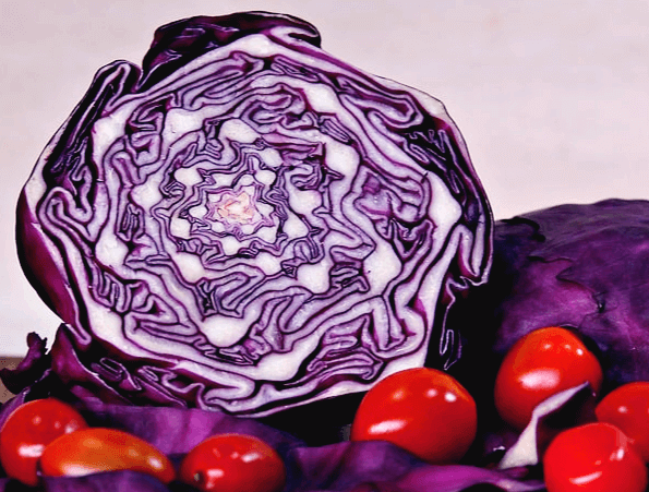 적양배추를 반으로 자른 단면으로 안은 보라색과 하얀색이 뒤섞였다. 앞에는 방울토마토 몇개가 있고 뒤에는 적양배추 한 통이 더 있다.