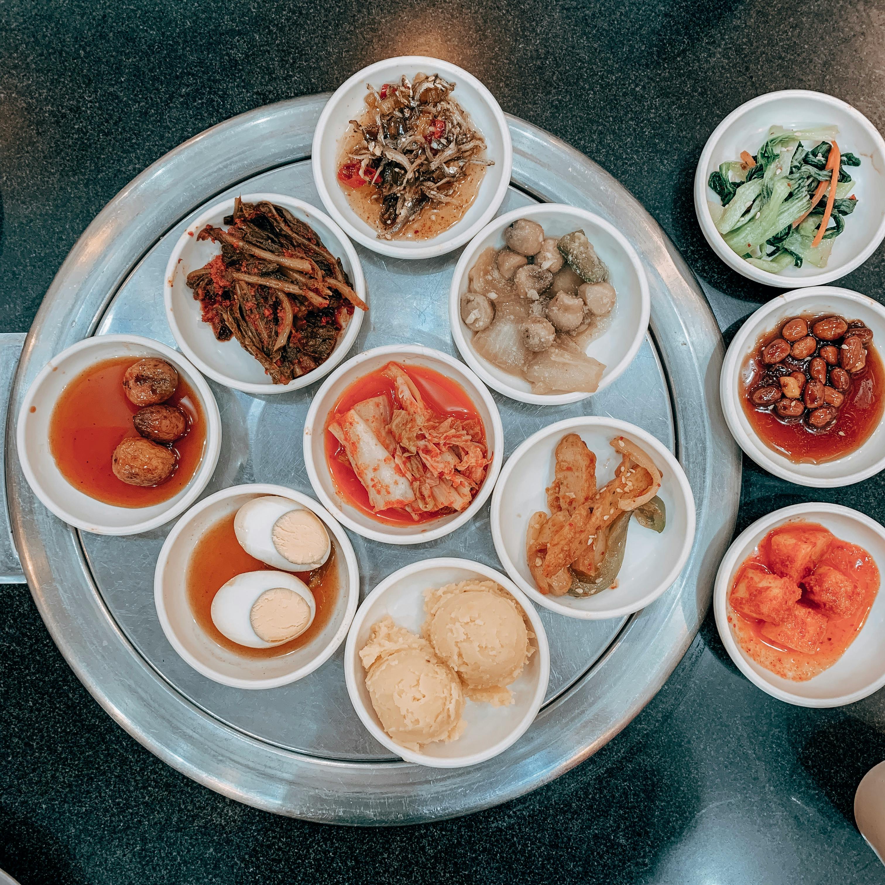 A top-down view of a circular tray filled with various Korean side dishes, or 'banchan', served in small white bowls, showcasing a colorful assortment of flavors.