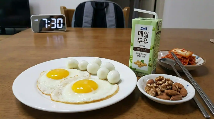 식탁 위 큰 접시에 계란 프라이, 메추리알 7개가 있고, 다른 접시에는 견과류, 또 다른 접시에는 김치가 담겨 있고, 두유 1팩이 놓여 있음