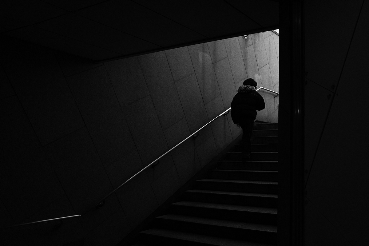 지하철 출구를 나가기 위해서 계단을 올라가고 있는 한 여성. 컬러가 사라진 흑백 사진이라 출구를 통해 살며시 들어오는 빛이 계단을 오르는 여성을 비춘다