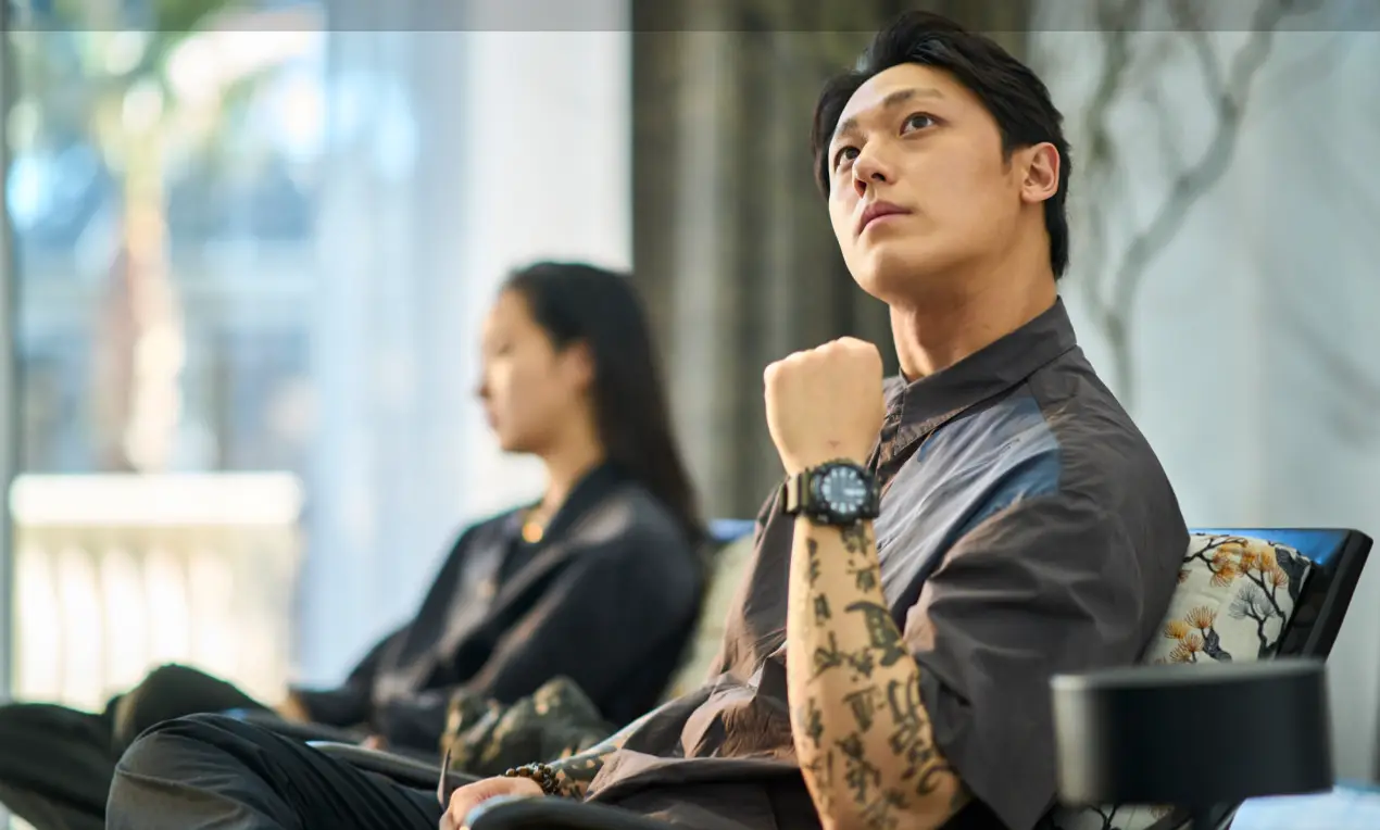 파묘- 회색옷을 입고 의자에 앉아 복잡하게 한문이 적힌 오른손을 팔걸이에 기대 올린채 위를 바라보는 이도현과 같이 앉아 있는 검은옷을 입은 이고은