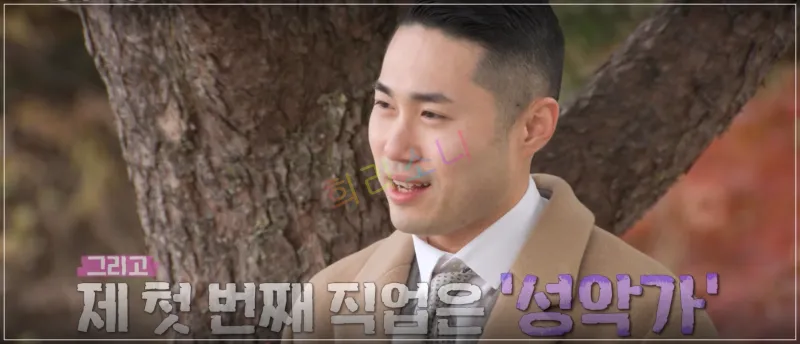 영수가 같은 장소에서 미소를 지으며 이야기하고 있다. 그는 같은 베이지색 코트를 입고 있으며&amp;#44; 가을 분위기의 배경이 보인다. 자막: 