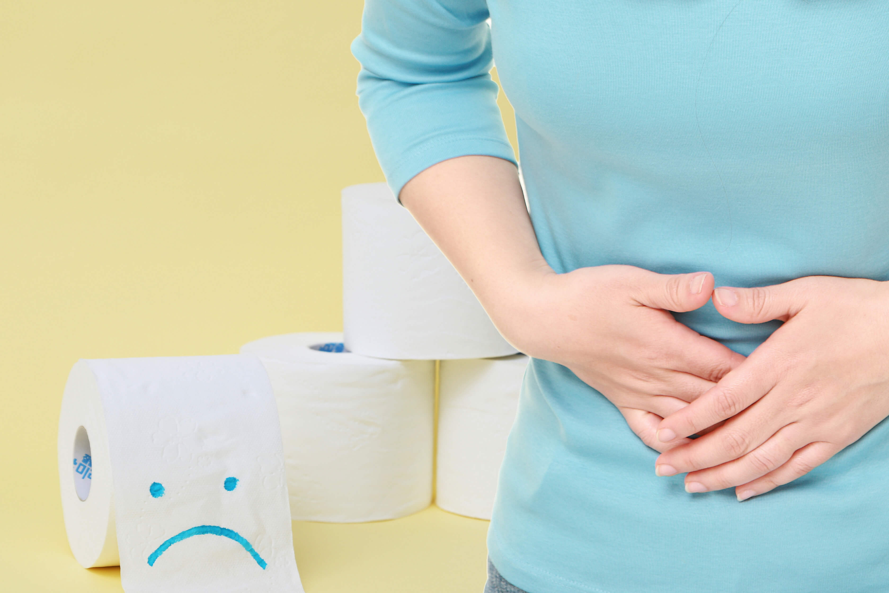 변비로 힘들어서 배를 움켜 쥐고 있는 여성과 그 뒤로 롤화장지가 놓여 있는 이미지