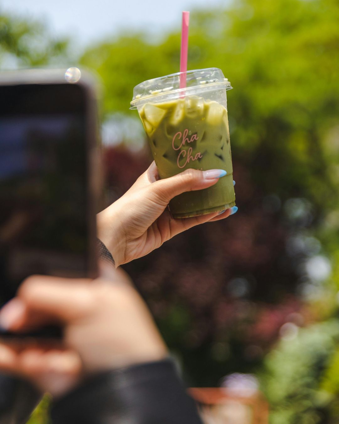 손에 들린 투명한 컵의 아이스 말차 라떼, 여름 햇살 아래 자연 배경 속 상큼한 분위기