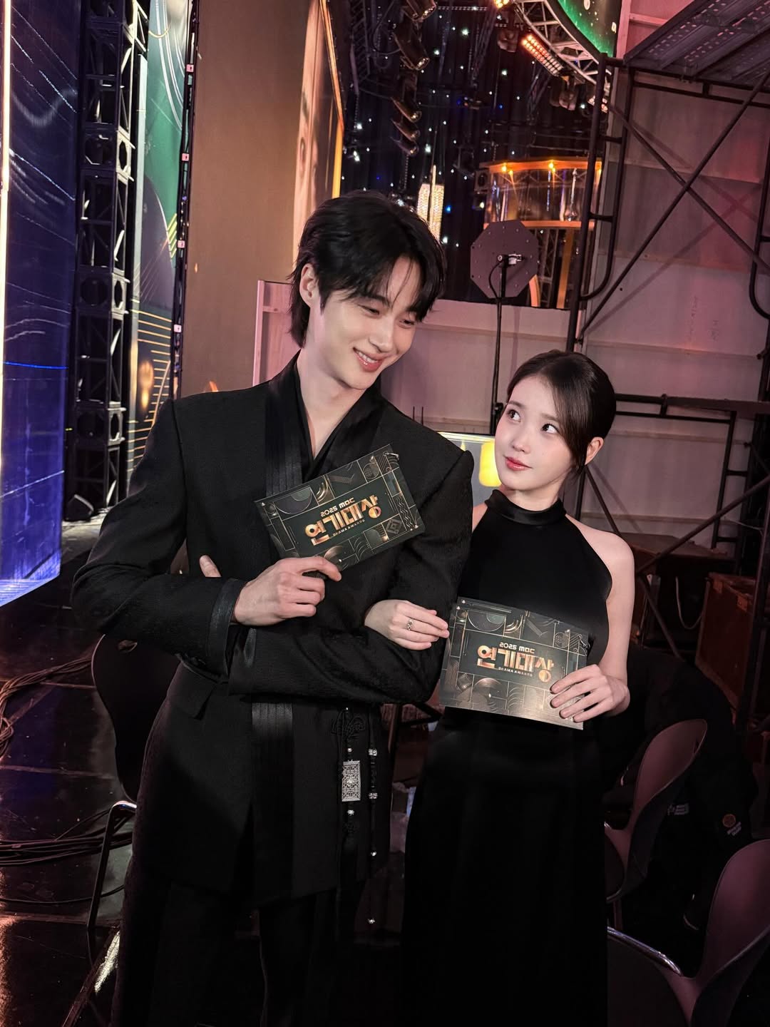 블랙 커플룩을 입고 2025 MBC 연기대상 무대에서 팔짱을 끼고 미소 짓는 아이유(성희주 역)와 변우석(이안대군 역)