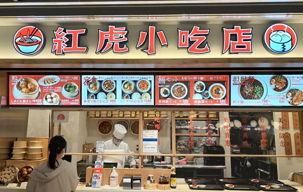 푸드코트내의 중국식당 정면 사진 메뉴가 가게위에 위치해 있고 점원이 밑에 서 있다.