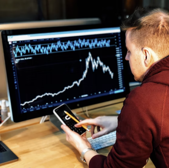 컴퓨터 모니터 화면에 차트가 보이며, 모니터 앞 책상에 않아있는 남성이 휴대폰을 보고 있다.