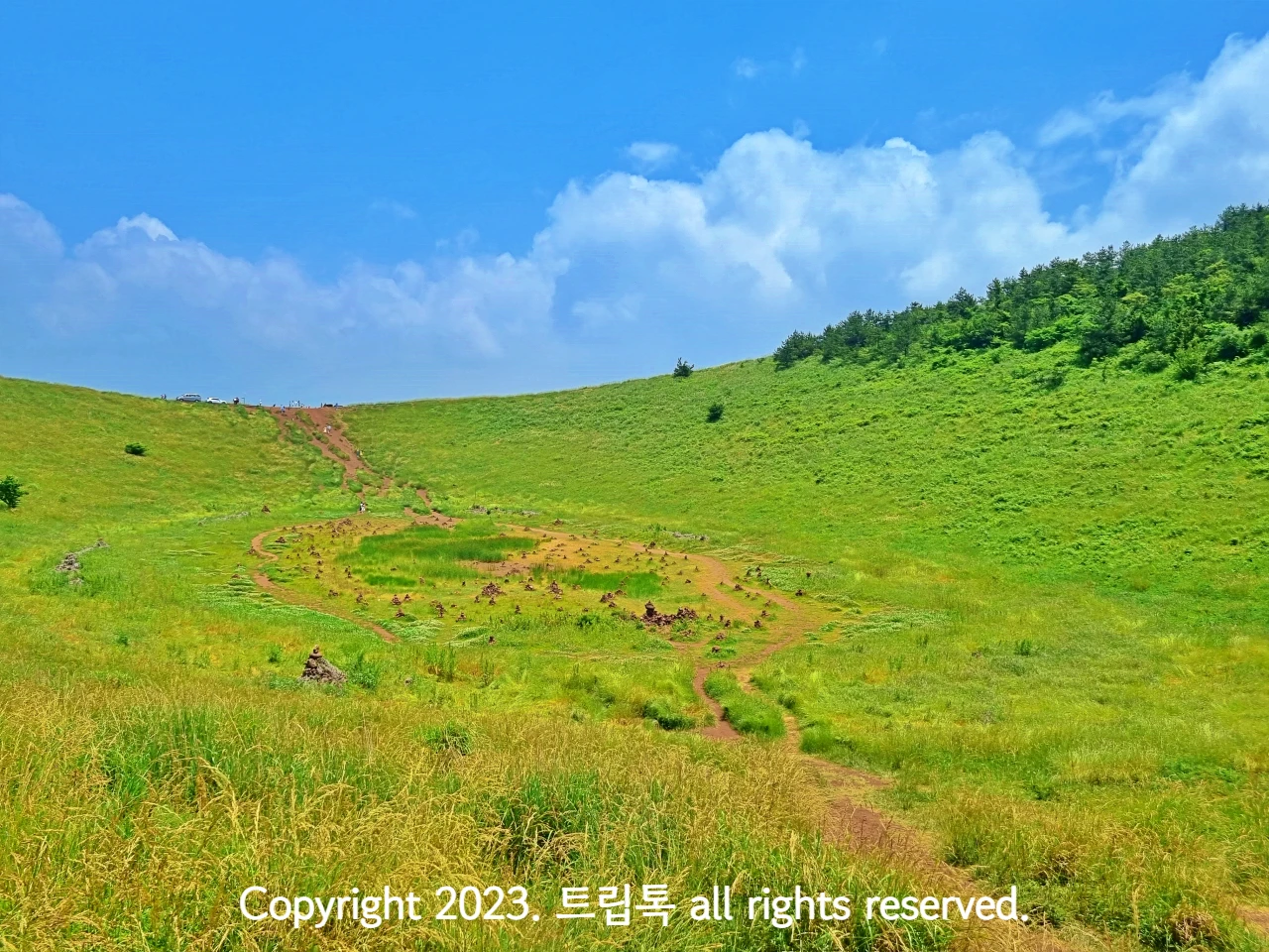 파란 하늘아래 움푹패인 연두빛 금오름의 분화구가 보인다. 가운에 동그란 흙길이 있다.