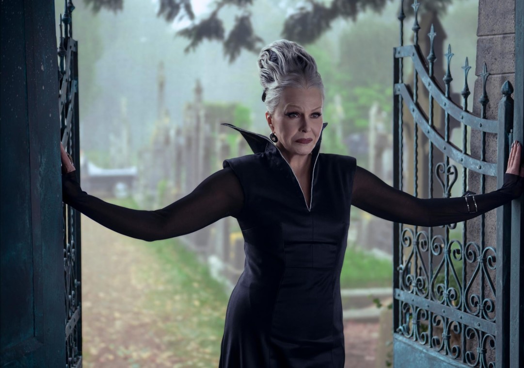 넷플릭스 웬즈데이 시즌2 신규 캐릭터 프럼프 할머니 역의 조안나 럼리 – 검은 드레스를 입고 묘지 문을 잡고 서 있는 장면