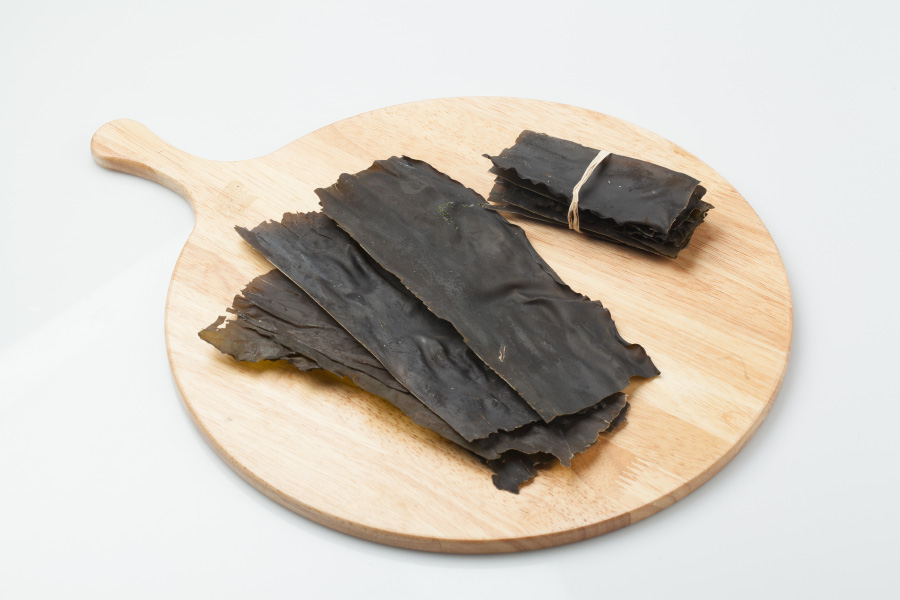 바다에서 채취를 한 다시마를 깨끗하게 세척한 후에 바싼 말린 후에 직사각형으로 잘라놓은 것을 하얀 바닥 위에 나무로 만든 동그란 깔판 위에 수북하게 놓아두고 찍은 사진