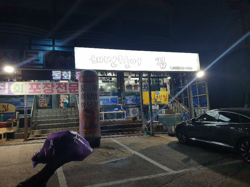 깜깜한 하늘 해변활어 집이라는 밝은 간판이 보인다. 그 옆 포장전문이라는 프프랜카드도 보인다.