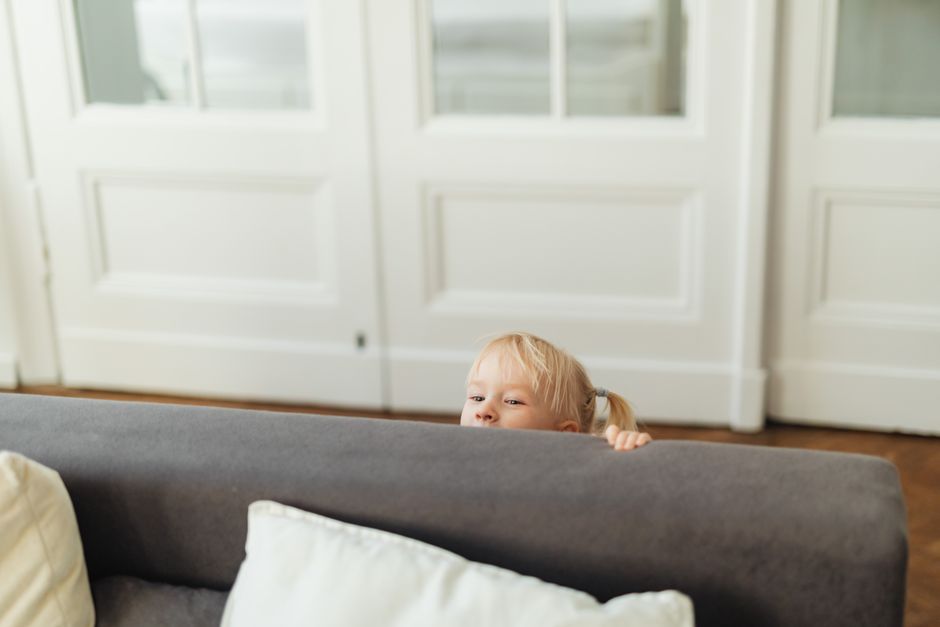 A blonde child playfully peeks over a gray sofa in a cozy living room setting.