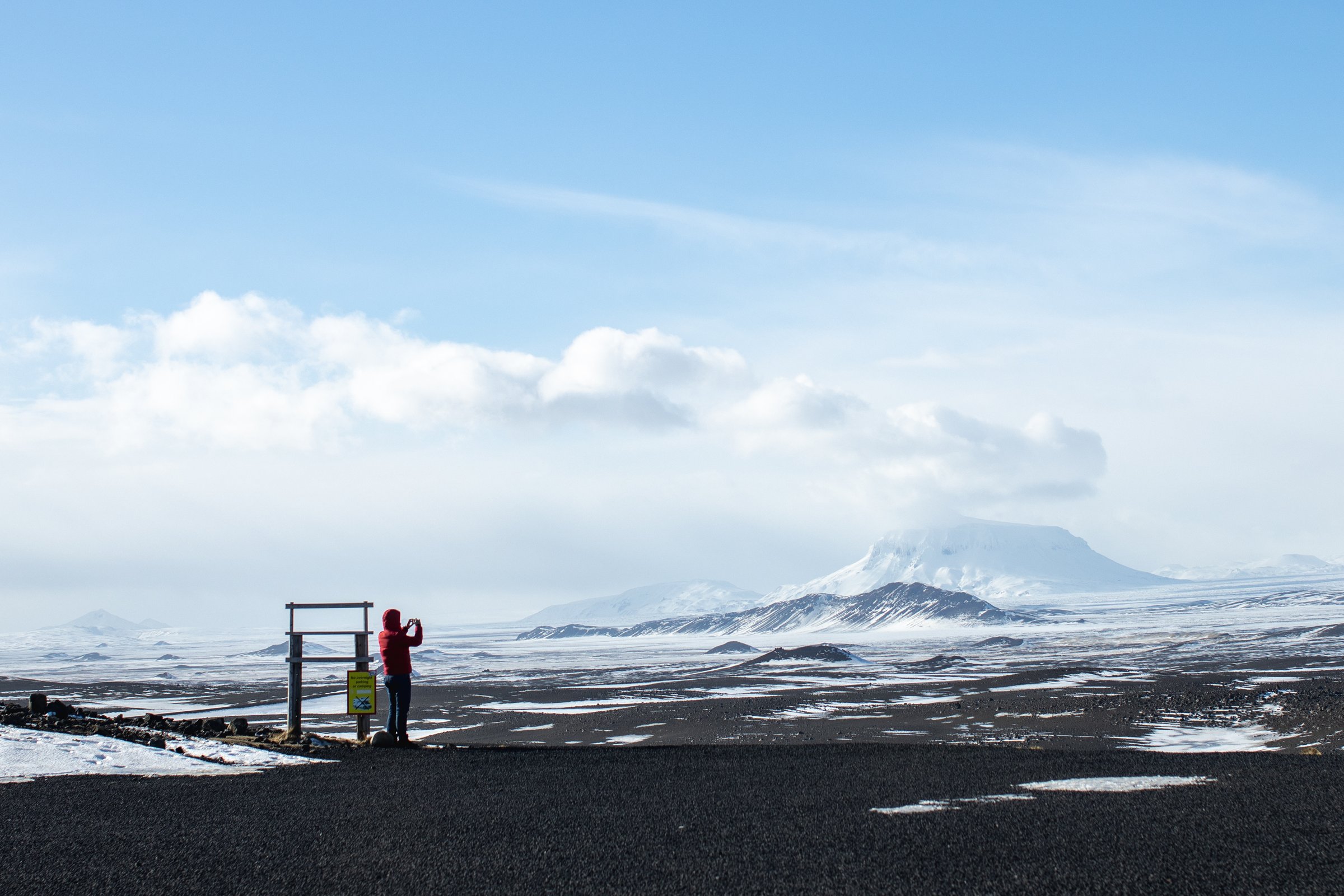 아이슬란드 화산 지형의 광활한 풍경입니다. 검은 화산재 지대와 부분적으로 눈이 덮인 평원이 펼쳐져 있고, 먼 배경에는 눈 덮인 산이 있습니다. 왼쪽 전경에는 빨간 재킷을 입은 사람이 조망 안내판 옆에 서서 스마트폰으로 풍경을 촬영하고 있습니다. 맑은 푸른 하늘과 흰 구름이 장엄한 풍경을 완성합니다. 이 사진은 아이슬란드의 극적인 자연 환경과 기후 조건을 잘 보여줍니다.