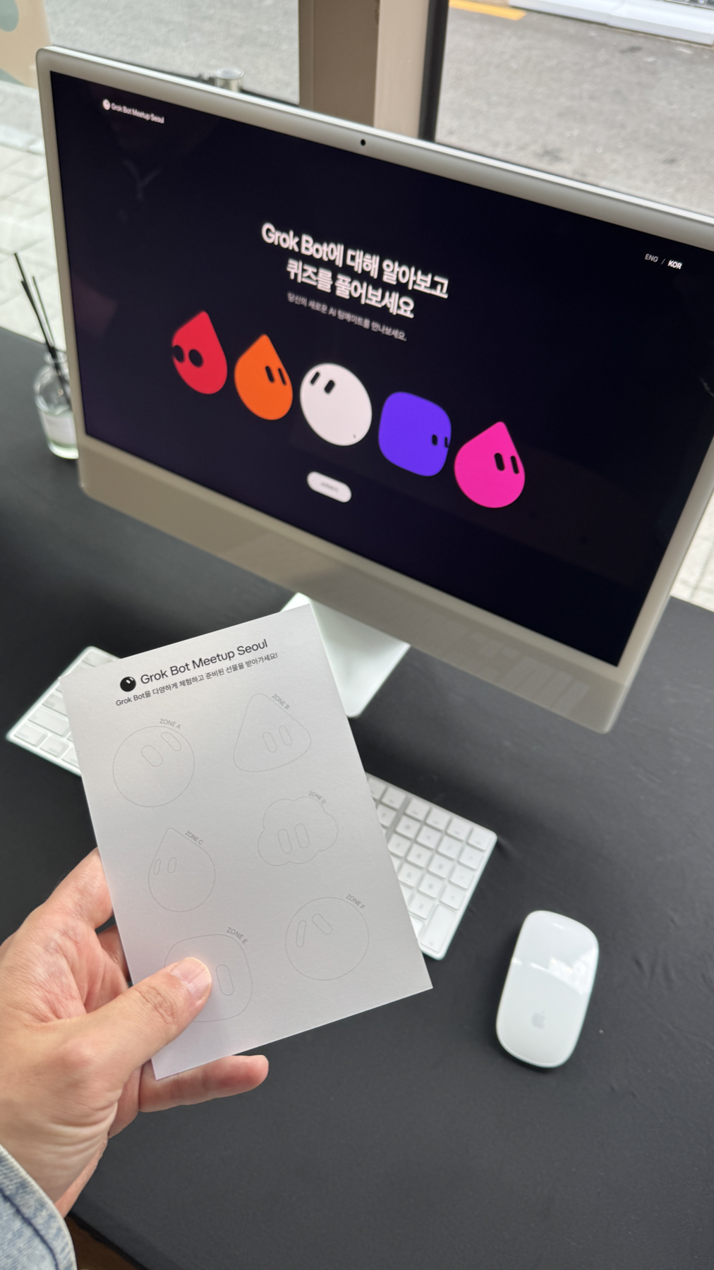 Grok Bot 퀴즈가 열린 iMac 앞에서 스탬프 카드를 손에 든 장면