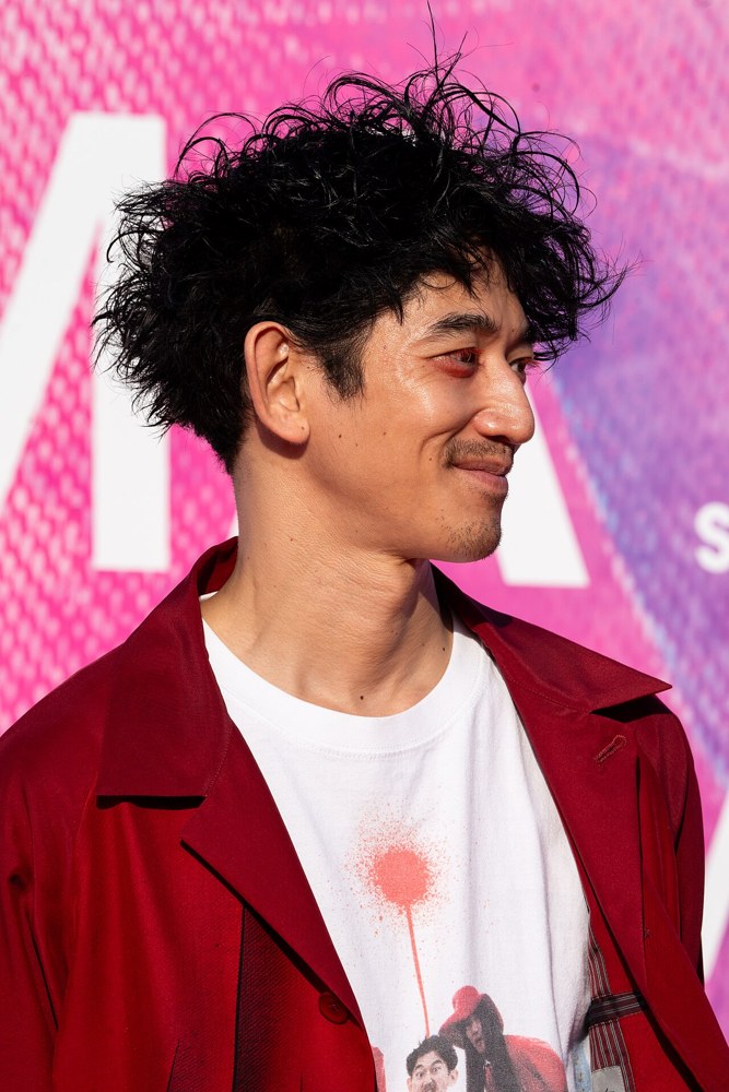 분홍색 배경 앞에서 붉은 재킷을 입고 옆을 보며 웃는 나가야마 에이타