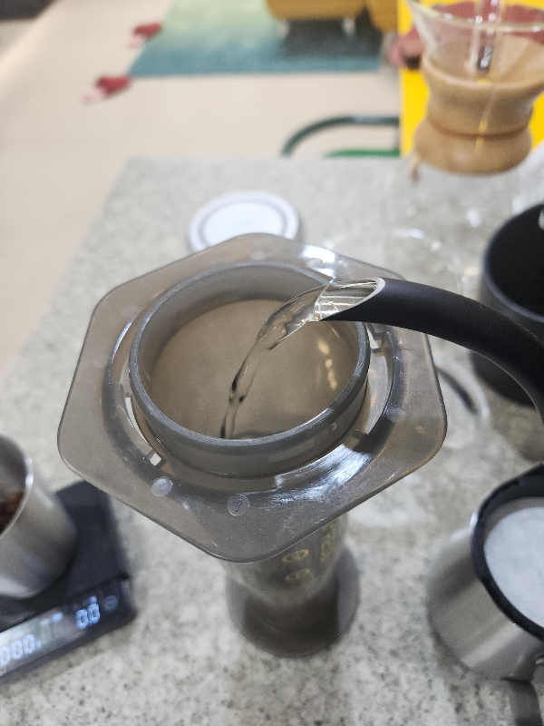 테이블 위에 역방향으로 놓인 에어로프레스에 예열을 위해 뜨거운 물을 붓고 있다.