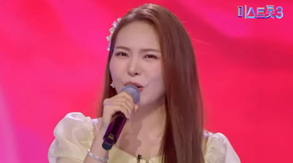 미스트롯3-
빨간배경 노란 드레스를 입고 긴머리를 늘어뜨린채 오른손으로 마이크를 잡고 노래하는 미스김