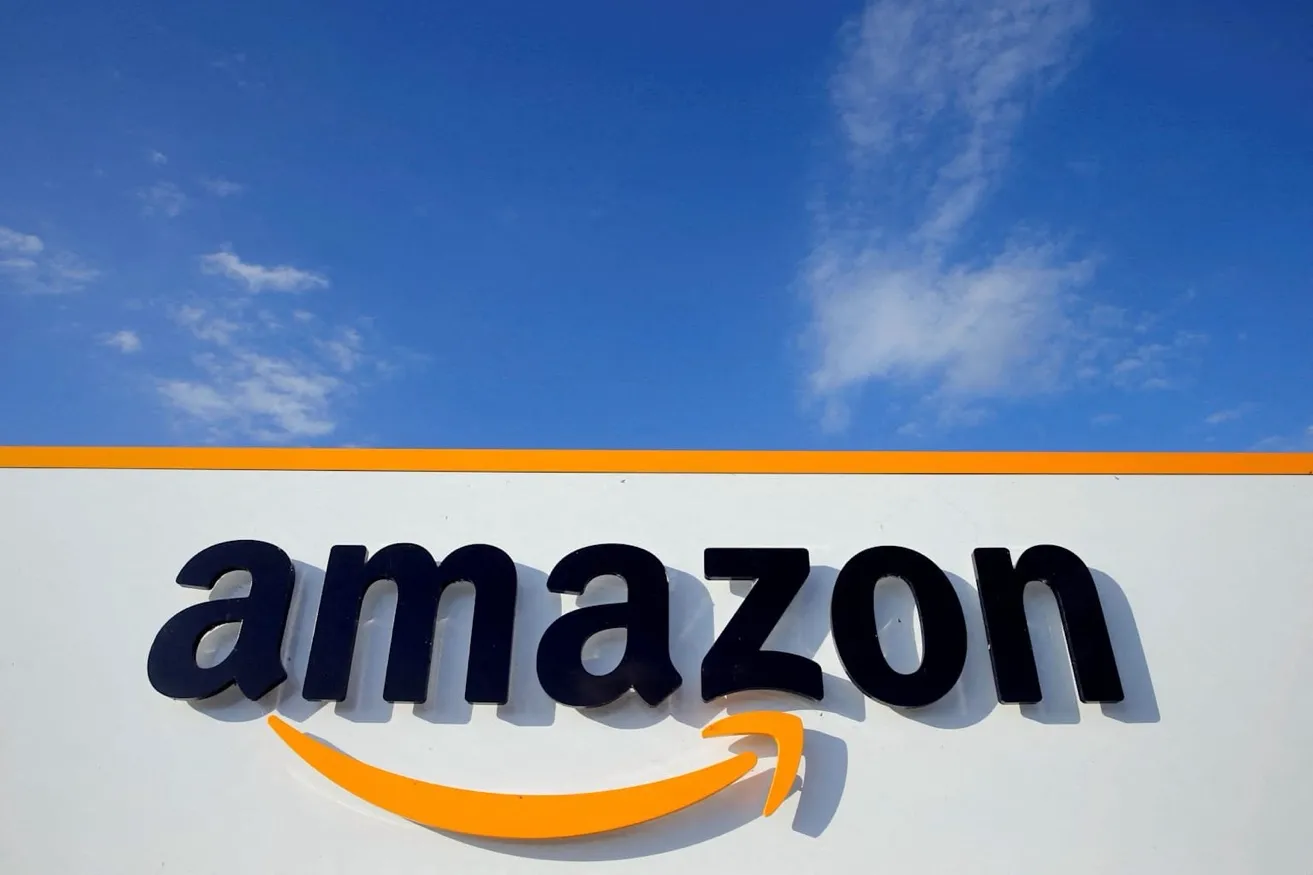 아마존 로고가 흰색 건물 외벽에 설치되어 있다. 로고는 소문자 검은색 글자로 'amazon'이라고 쓰여 있으며, 아래에 주황색 화살표 모양의 곡선이 'a'에서 'z'까지 연결되어 미소 짓는 모양을 형성하고 있다. 배경은 맑은 파란 하늘이며 건물 상단에 주황색 띠가 가로로 길게 이어져 있다. 전형적인 아마존 물류센터나 사무실 건물의 외관이다.
