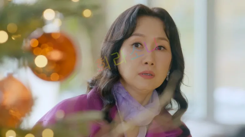 장미애(배해선)가 약간 놀란 듯한 표정으로 어딘가를 바라보고 있다. 그녀는 보라색 코트를 입고 있으며&amp;#44; 목에는 연보라색 스카프를 두르고 있다. 앞쪽에는 크리스마스 트리 장식이 보이며&amp;#44; 오렌지색 오너먼트가 빛을 반사하고 있다. 배경은 창문을 통해 자연광이 들어오는 밝은 실내 공간이다.