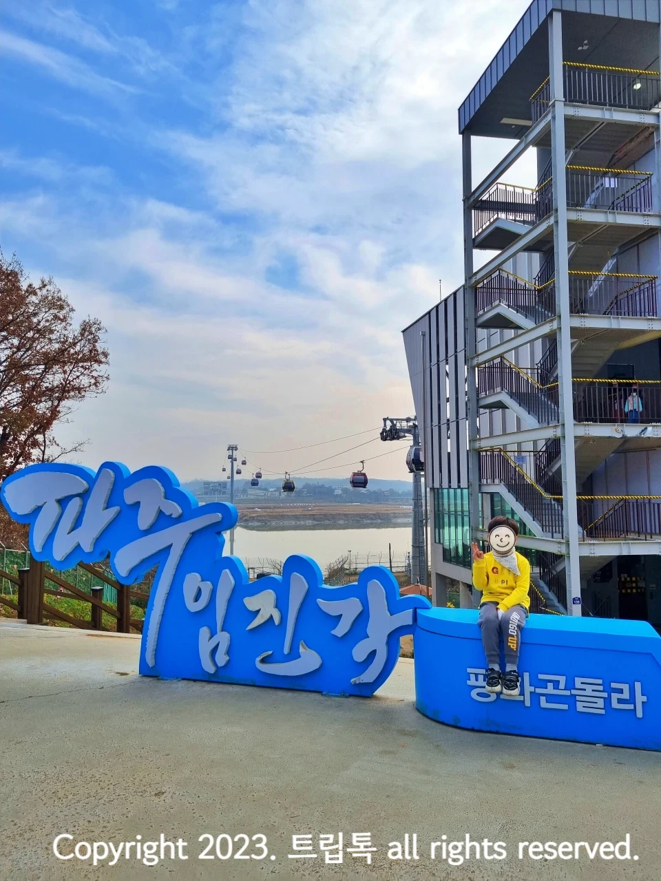 파란색 배경의 하얀 글씨로 파주임진각 평화곤돌라라고 쓰인 커다란 조형물이 있고 그 위에 한 아이가 앉아있다. 그 뒤오 전망대와 케이블카가 보인다.