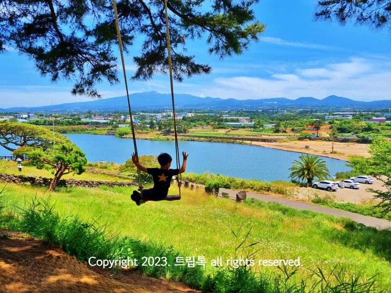 한라산과 수산 저수지가 보이는 넓은 곳에서 한 아이가 양손 만세를 하고 하늘그네를 타고 있는 뒷모습