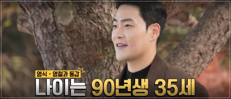상철이 야외에서 나무 앞에서 인터뷰를 하고 있다. 그는 검은색 이너웨어와 갈색 재킷을 입고 있으며&amp;#44; 짧은 머리에 깔끔한 인상을 준다. 배경은 가을 분위기의 풍경이다. 자막: 