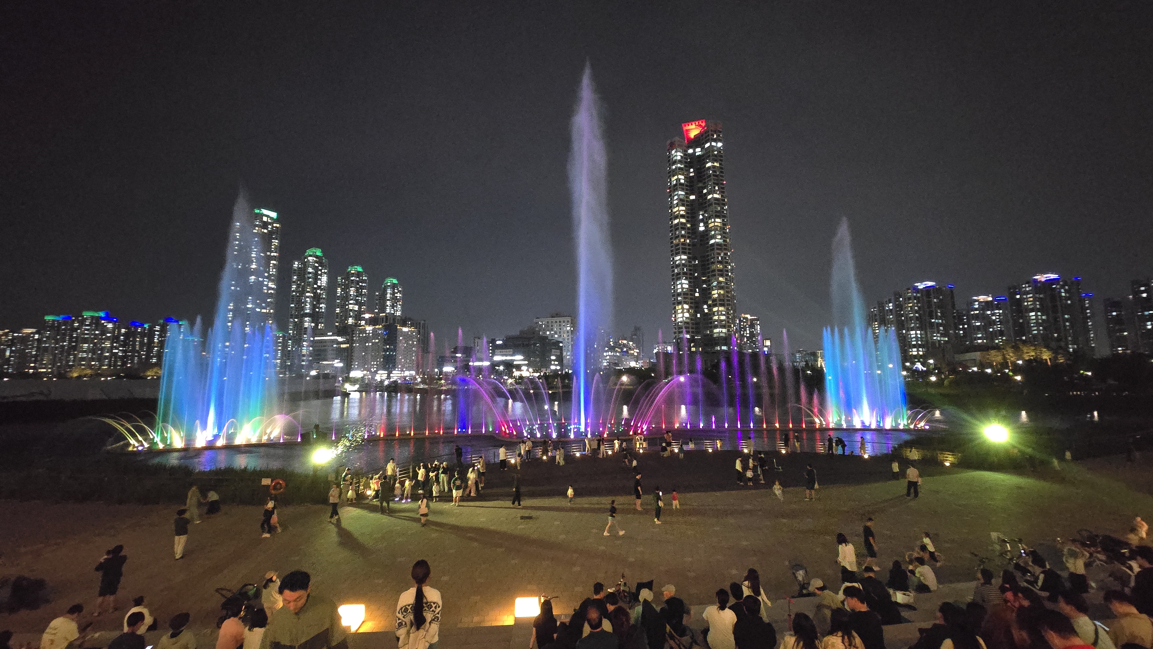 청라호수공원 음악분수 공연을 관람 중인 시민들, 형형색색의 조명과 고층 빌딩 야경이 어우러진 전경