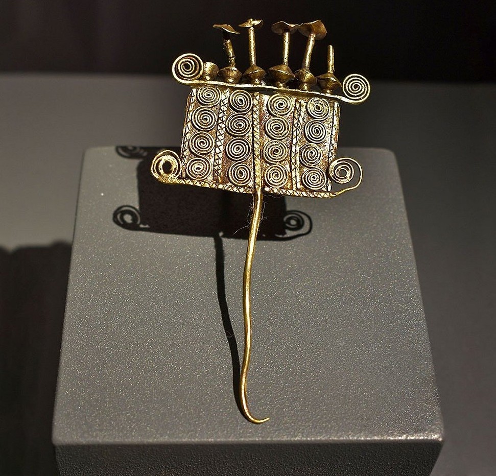 금으로 된 핀 장식이 진열대에 놓여 있다. 상단에는 나선형 문양이 3열로 반복되며, 작은 꽃 모양 장식이 위에 장착되어 있고, 긴 바늘 형태의 핀이 아래로 뻗어 있다.