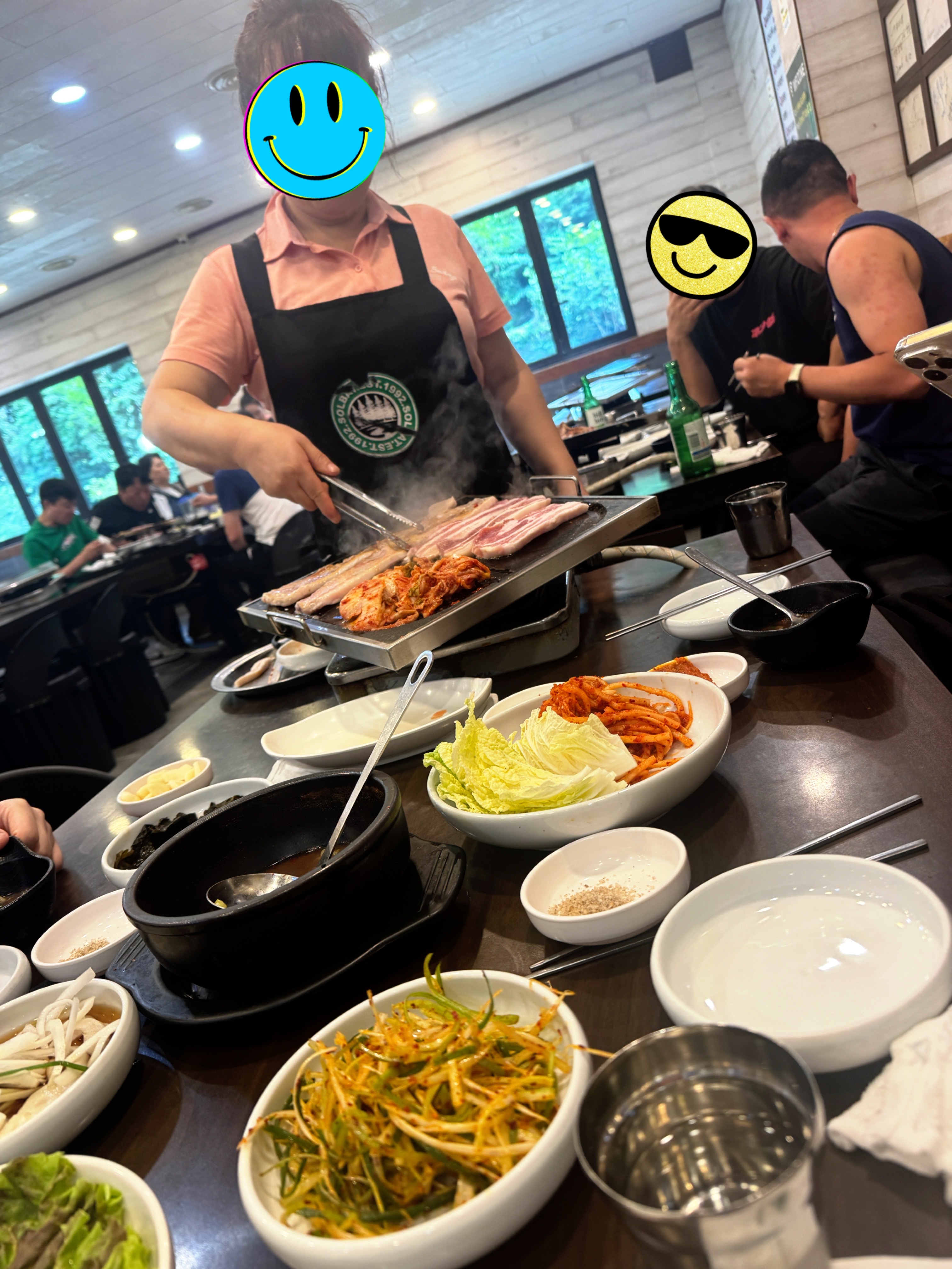 솔밭삼겹살에서 이모님이 불판 위 삼겹살과 김치를 정성껏 구워주시는 모습. 주변 밑반찬과 손님들 모습도 함께 담겨 있다.