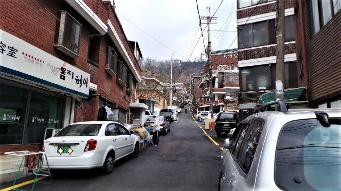 골목 양쪽에 븕은 벽돌집들로 이어지는 산동네 골목풍경, 양쪽에 주차된 차량들이 많아요,