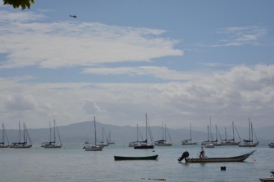 브라질 플로리아노폴리스 해안가에 정박해 있는 여러 대의 요트와 돛단배들, 멀리 보이는 산맥과 맑은 하늘 위로 날아가는 헬리콥터