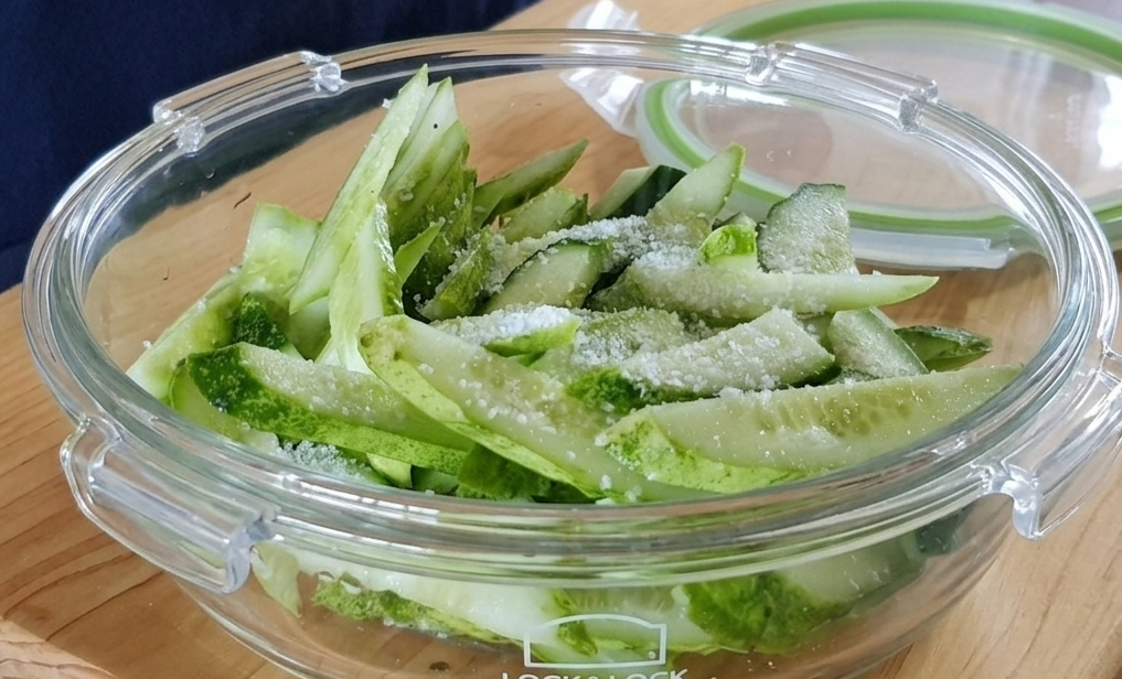 투명한 유리 밀폐 용기에 담긴 손질된 오이와 그 위에 뿌려진 굵은 소금, 오이무침 절이기 과정.