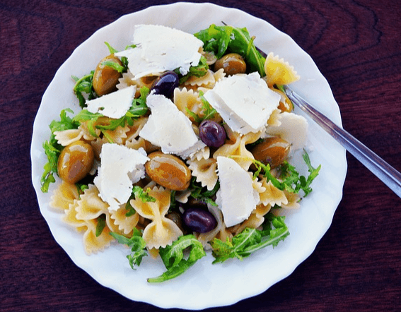 하얀 접시에 나비 모양의 파스타와 초록색 루꼴라들, 그리고 하얀 치즈덩이들이 가득하다.