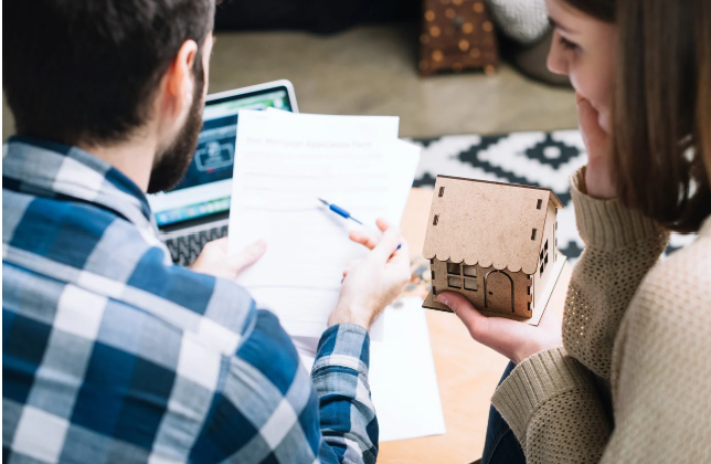 보험 계약서를 검토 중인 젊은 부부와 작은 집 모형을 들고 있는 여성의 모습