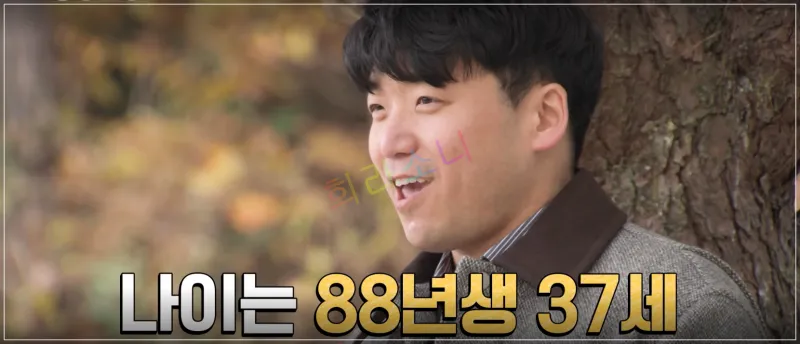 영호가 야외에서 나무 옆에 서서 활짝 웃고 있다. 그는 갈색 계열의 체크무늬 재킷을 입고 있으며&amp;#44; 가을 분위기의 배경이 펼쳐져 있다. 자막: 